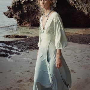 May include: A light green dress with a deep V-neck and long sleeves. The dress is paired with a matching head wrap and turquoise jewelry. The model is wearing studded ankle boots. The background is a beach with rocks and the ocean.