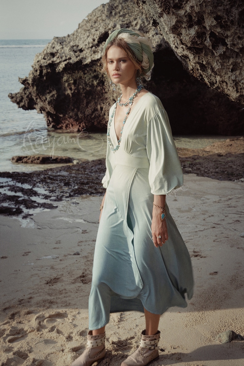 May include: A light green dress with a deep V-neck and long sleeves. The dress is paired with a matching head wrap and turquoise jewelry. The model is wearing studded ankle boots. The background is a beach with rocks and the ocean.