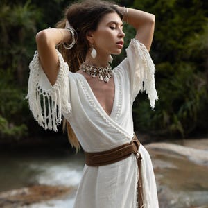 Puede incluir: Un vestido blanco cruzado con mangas con flecos y un cintur&oacute;n marr&oacute;n. El vestido tiene un cuello en V y una falda fluida. La modelo lleva un collar y pendientes. El fondo es un entorno natural con agua y vegetaci&oacute;n.