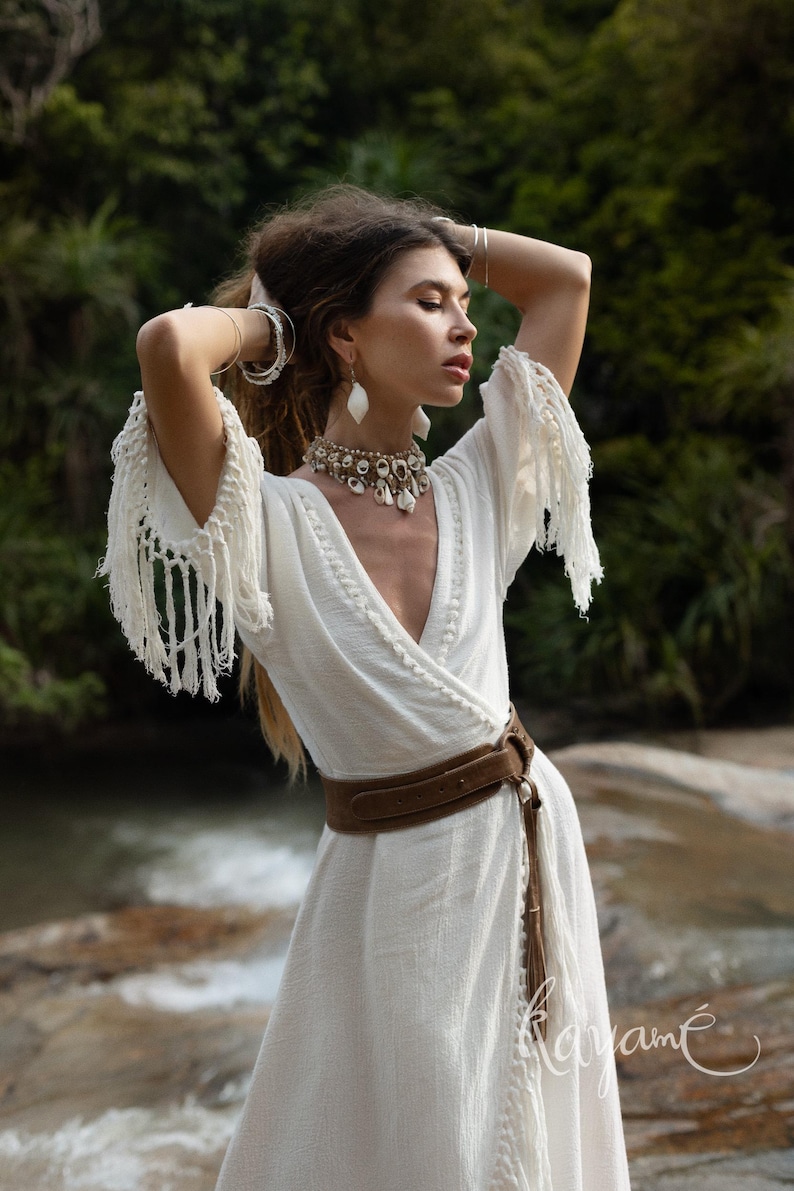 Puede incluir: Un vestido blanco cruzado con mangas con flecos y un cintur&oacute;n marr&oacute;n. El vestido tiene un cuello en V y una falda fluida. La modelo lleva un collar y pendientes. El fondo es un entorno natural con agua y vegetaci&oacute;n.
