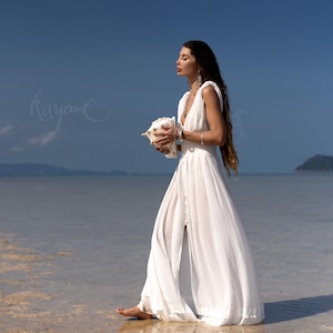 May include: A woman in a white, flowing dress walks in shallow water, holding a large seashell. The dress has a deep V-neck and a button-down front. The background features a clear blue sky and ocean.