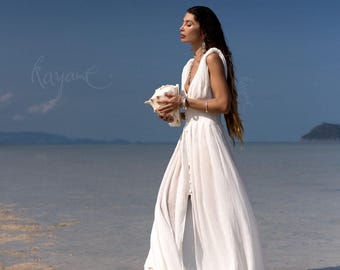 May include: A woman in a white, flowing dress walks in shallow water, holding a large seashell. The dress has a deep V-neck and a button-down front. The background features a clear blue sky and ocean.