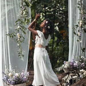 Puede incluir: Una mujer con un vestido blanco y una corona de flores se encuentra en un entorno boscoso. Est&aacute; rodeada de vegetaci&oacute;n y flores, y la luz del sol se filtra a trav&eacute;s de los &aacute;rboles.