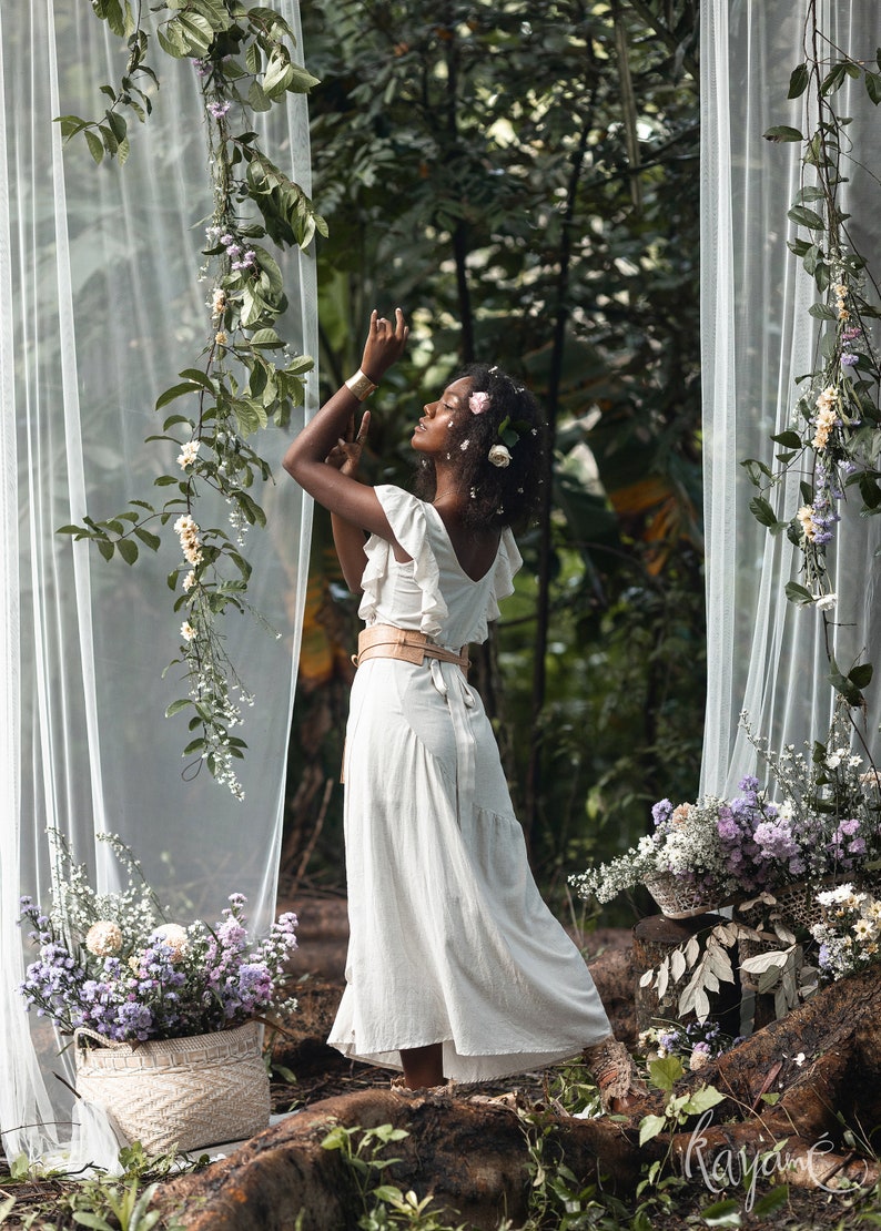 Puede incluir: Una mujer con un vestido blanco y una corona de flores se encuentra en un entorno boscoso. Est&aacute; rodeada de vegetaci&oacute;n y flores, y la luz del sol se filtra a trav&eacute;s de los &aacute;rboles.