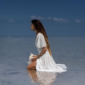 May include: A woman in a white dress kneels in shallow water, holding a large seashell. The dress has flowing sleeves and a long skirt. The background is a clear blue sky and calm water. The image has a serene and ethereal quality.