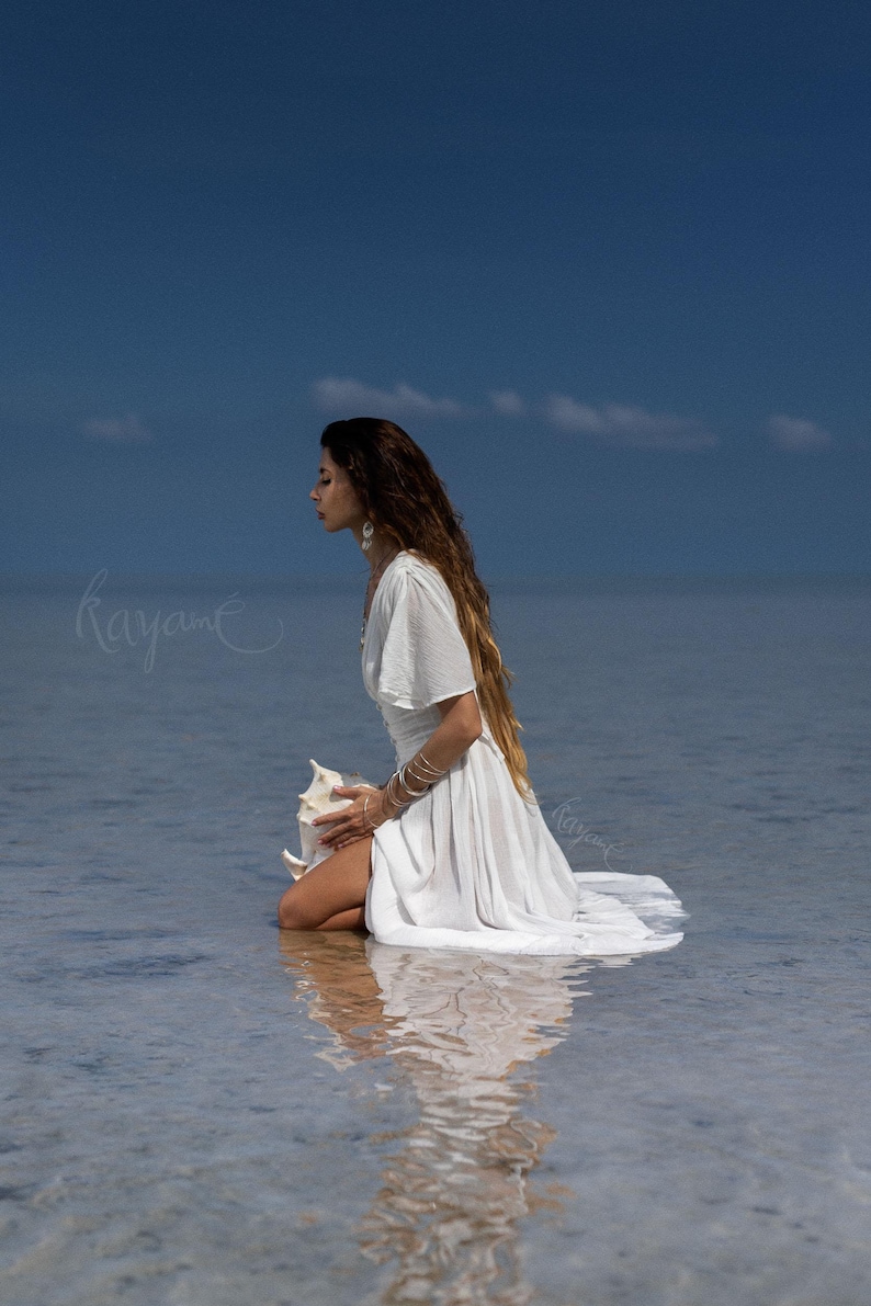 May include: A woman in a white dress kneels in shallow water, holding a large seashell. The dress has flowing sleeves and a long skirt. The background is a clear blue sky and calm water. The image has a serene and ethereal quality.