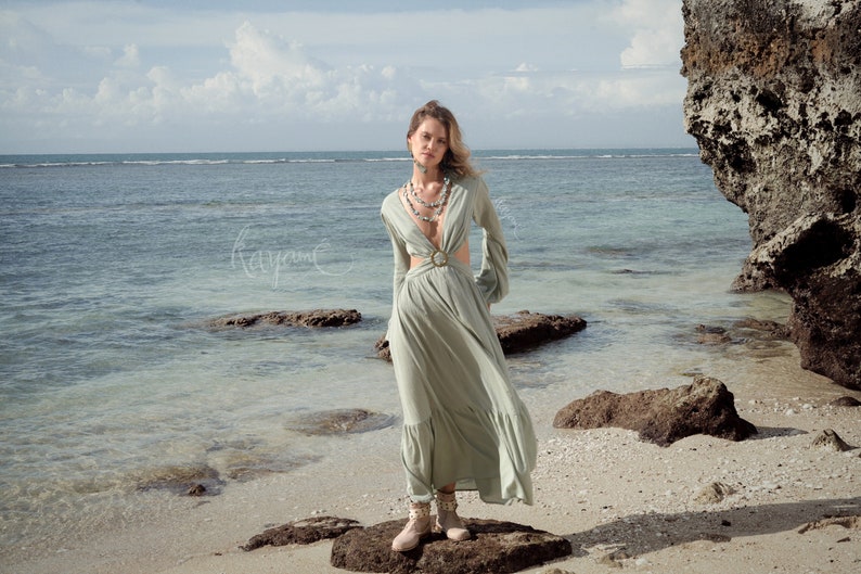 Puede incluir: Una mujer con un vestido maxi verde claro con un dise&ntilde;o de corte y un cintur&oacute;n. Est&aacute; de pie sobre una roca en una playa con el oc&eacute;ano de fondo.