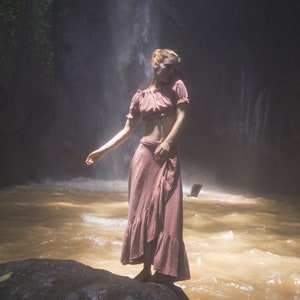 May include: A woman wearing a pink crop top and a long pink skirt stands in front of a waterfall. The water is flowing over rocks and creating a mist.