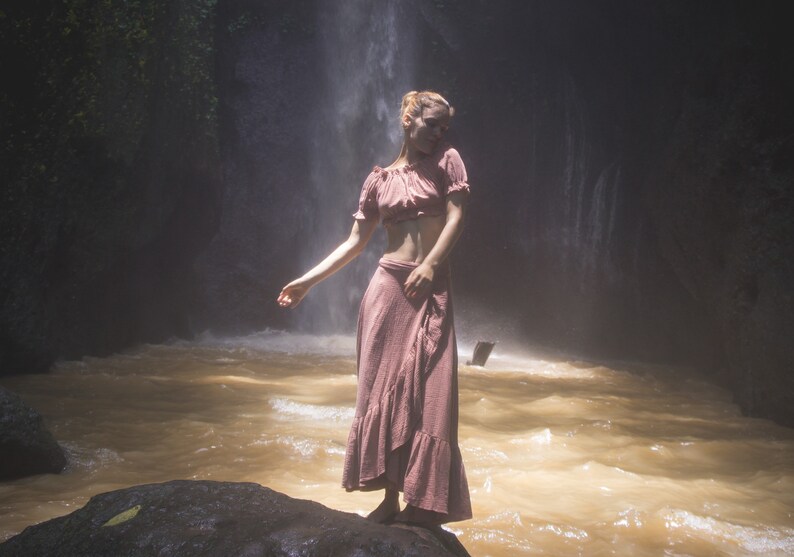 May include: A woman wearing a pink crop top and a long pink skirt stands in front of a waterfall. The water is flowing over rocks and creating a mist.