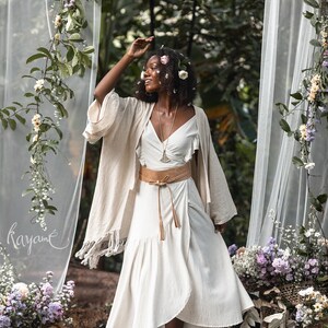 Puede incluir: Una mujer con un vestido blanco con un cintur&oacute;n marr&oacute;n y un kimono beige. Est&aacute; de pie en un entorno de bosque con flores y vegetaci&oacute;n a su alrededor. La palabra "rayam&eacute;" est&aacute; escrita en una cortina blanca transparente detr&aacute;s de ella.