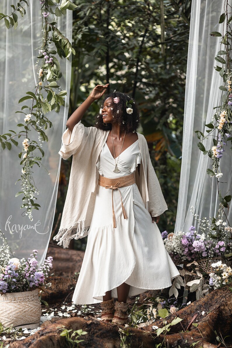 Puede incluir: Una mujer con un vestido blanco con un cintur&oacute;n marr&oacute;n y un kimono beige. Est&aacute; de pie en un entorno de bosque con flores y vegetaci&oacute;n a su alrededor. La palabra "rayam&eacute;" est&aacute; escrita en una cortina blanca transparente detr&aacute;s de ella.