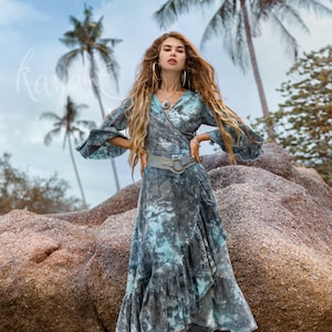 May include: A woman wearing a blue and white tie-dye wrap dress with a brown leather belt and white cowboy boots. She is standing on a large rock with palm trees in the background.