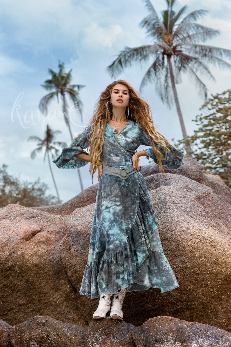 May include: A woman wearing a blue and white tie-dye wrap dress with a brown leather belt and white cowboy boots. She is standing on a large rock with palm trees in the background.
