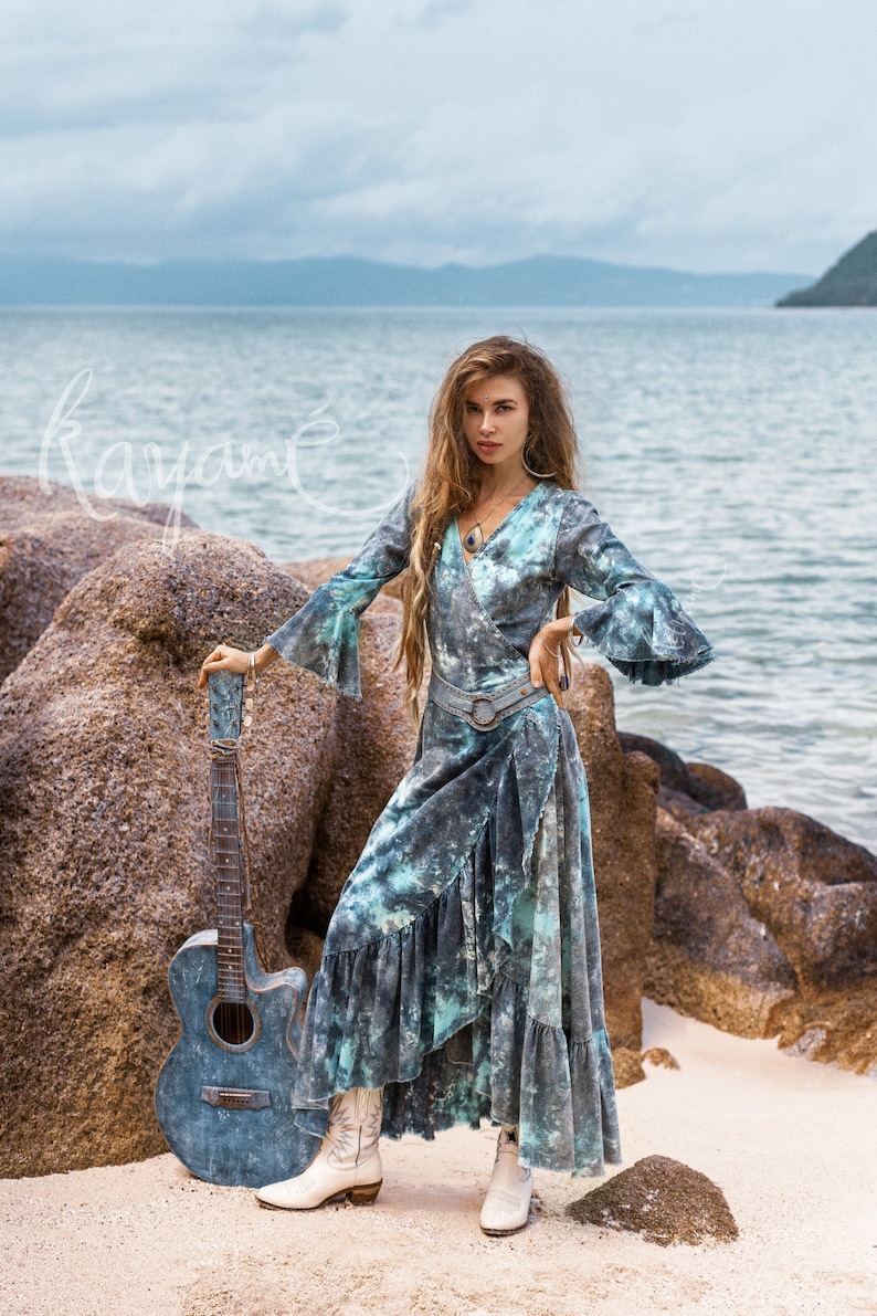 May include: A woman wearing a blue and green tie-dye wrap dress with a silver belt and white cowboy boots. She is standing on a beach with rocks and a guitar beside her.