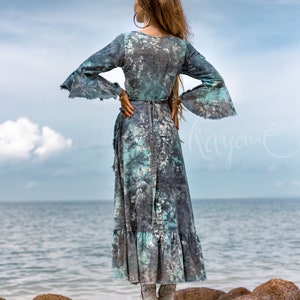 May include: A woman wearing a long, flowing blue and white tie-dye dress with a ruffled hem. She is standing on a rocky beach with the sea in the background.