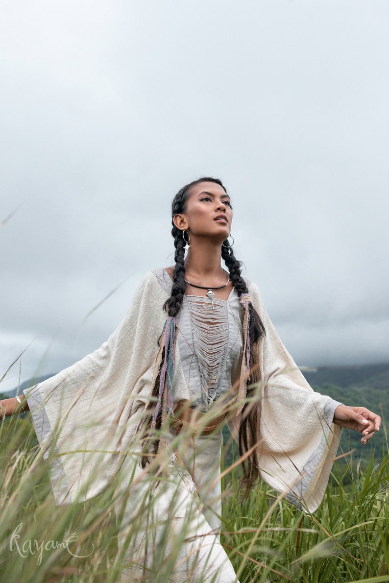 May include: A woman with long black braids wears a white, loose-fitting top with a crocheted design and a silver necklace. She stands in a field of tall grass, looking up at the sky.
