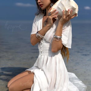 May include: A woman in a white dress kneels near the water, holding a large conch shell. She wears silver bracelets and jewelry. The dress has a buttoned bodice and flowing skirt. The background is the ocean and a blue sky.