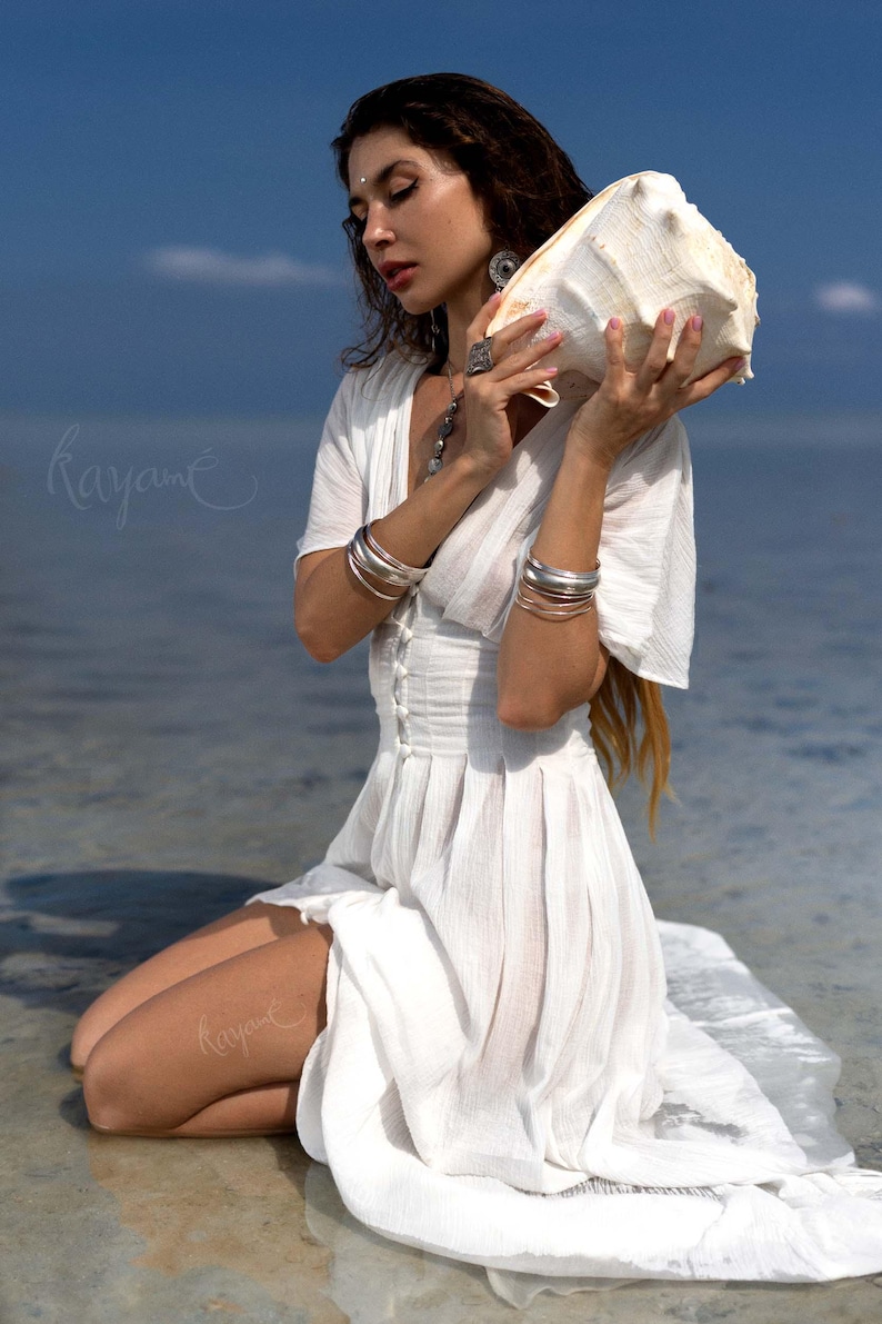May include: A woman in a white dress kneels near the water, holding a large conch shell. She wears silver bracelets and jewelry. The dress has a buttoned bodice and flowing skirt. The background is the ocean and a blue sky.
