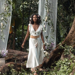 Puede incluir: Una mujer con un vestido blanco con un cintur&oacute;n marr&oacute;n y una corona de flores camina por un exuberante entorno de bosque verde. El vestido tiene un escote en V y mangas con volantes. La mujer est&aacute; rodeada de vegetaci&oacute;n y flores.
