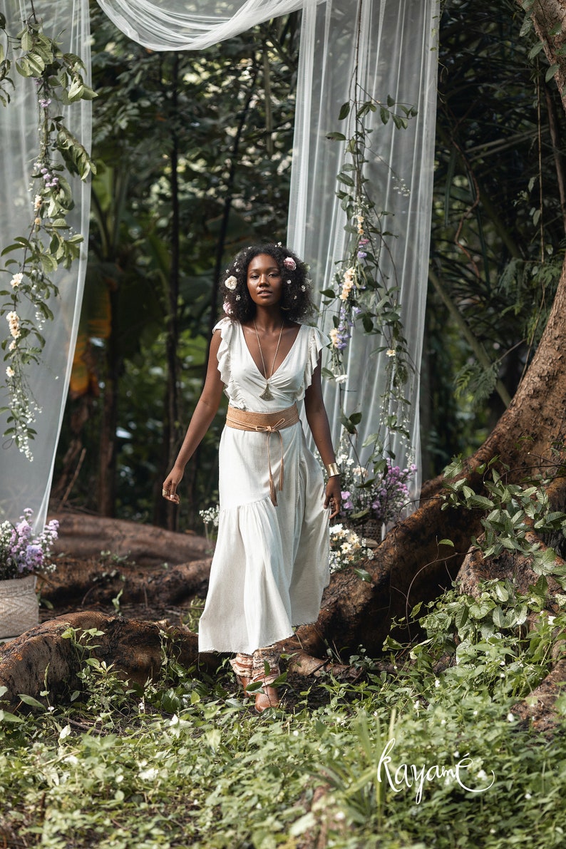Puede incluir: Una mujer con un vestido blanco con un cintur&oacute;n marr&oacute;n y una corona de flores camina por un exuberante entorno de bosque verde. El vestido tiene un escote en V y mangas con volantes. La mujer est&aacute; rodeada de vegetaci&oacute;n y flores.