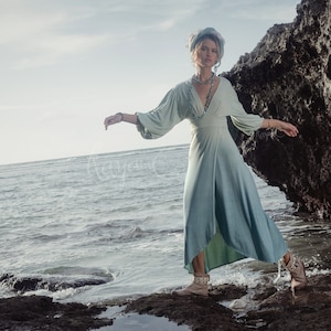 May include: A flowing, light blue dress with long sleeves and a V-neck. The dress is worn with a matching headscarf and jewelry. The model is standing on rocks near the ocean. The dress has a split on the side.