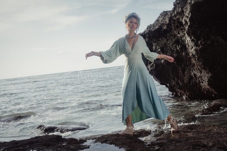 May include: A flowing, light blue dress with long sleeves and a V-neck. The dress is worn with a matching headscarf and jewelry. The model is standing on rocks near the ocean. The dress has a split on the side.