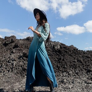 Puede incluir: Un vestido largo de manga larga con un degradado de color que va desde el verde salvia claro en la parte superior hasta un azul verdoso profundo en el dobladillo. El vestido tiene un escote en V profundo, una abertura alta y botas oscuras.