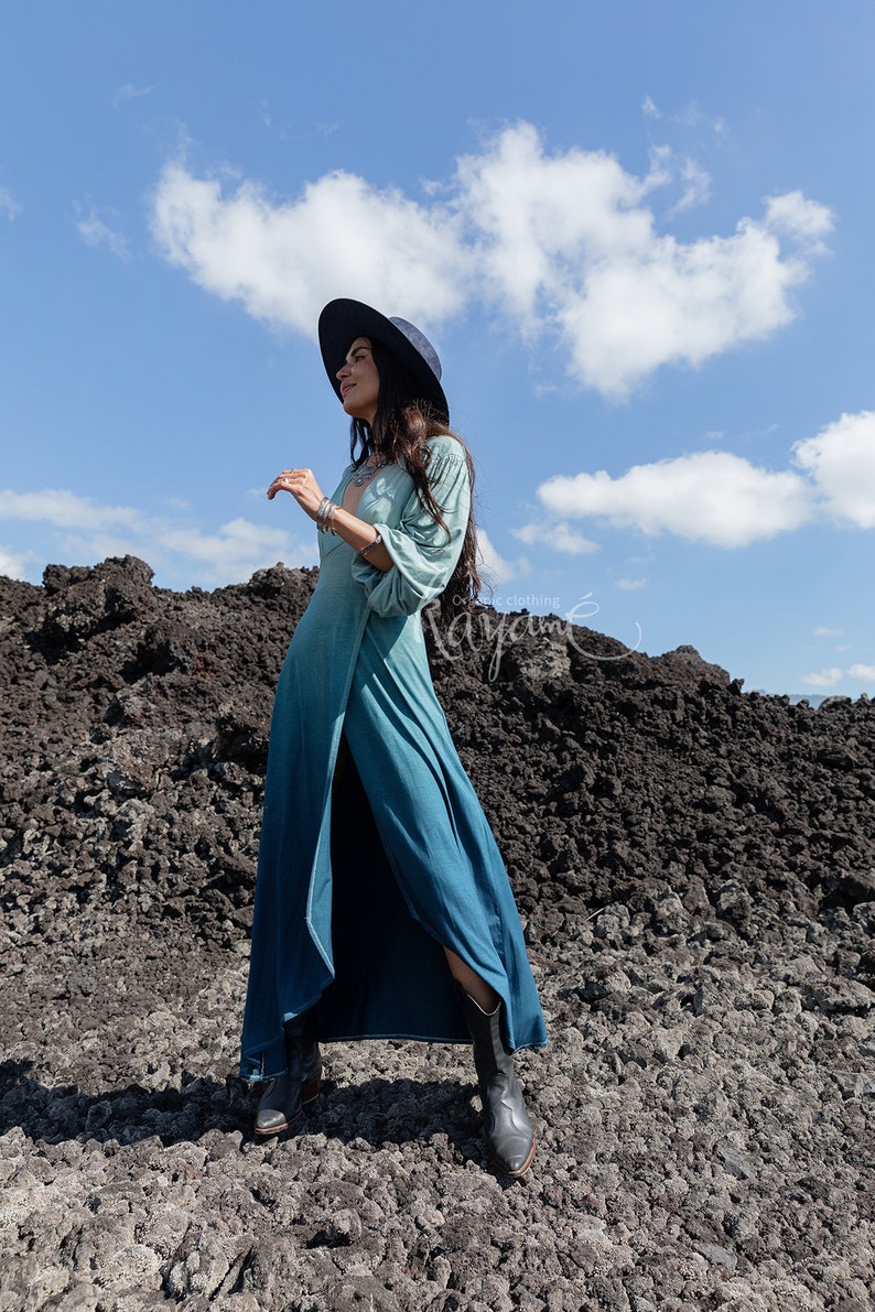 Puede incluir: Un vestido largo de manga larga con un degradado de color que va desde el verde salvia claro en la parte superior hasta un azul verdoso profundo en el dobladillo. El vestido tiene un escote en V profundo, una abertura alta y botas oscuras.