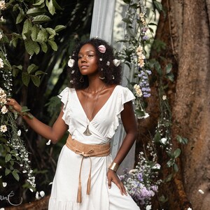 Puede incluir: Una mujer con un vestido blanco, un cintur&oacute;n de cuero marr&oacute;n y un collar de plata se encuentra en un jard&iacute;n. Lleva flores en el pelo y est&aacute; rodeada de vegetaci&oacute;n.