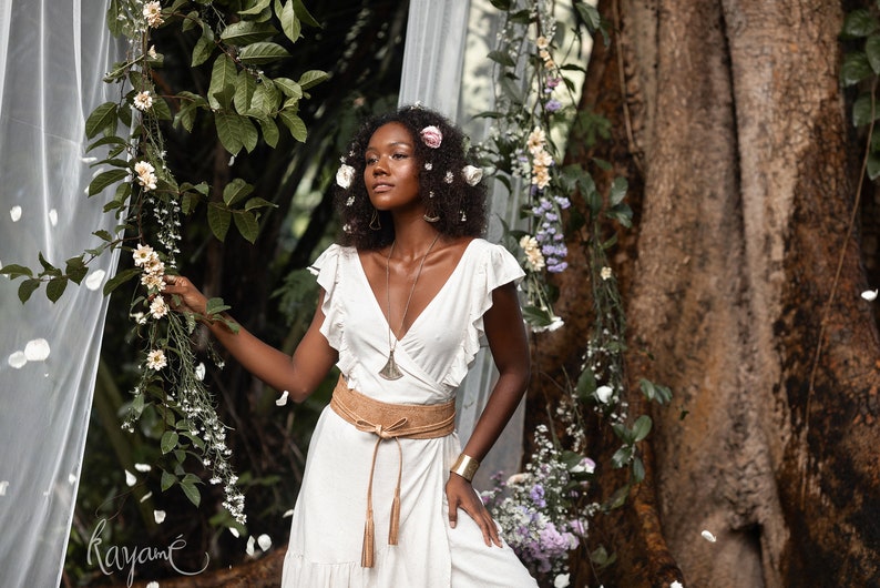 Puede incluir: Una mujer con un vestido blanco, un cintur&oacute;n de cuero marr&oacute;n y un collar de plata se encuentra en un jard&iacute;n. Lleva flores en el pelo y est&aacute; rodeada de vegetaci&oacute;n.