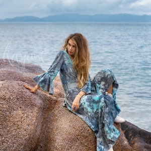May include: A woman wearing a blue and white tie-dye maxi dress with bell sleeves sits on a large rock by the ocean. She is wearing white boots and has long, wavy hair.
