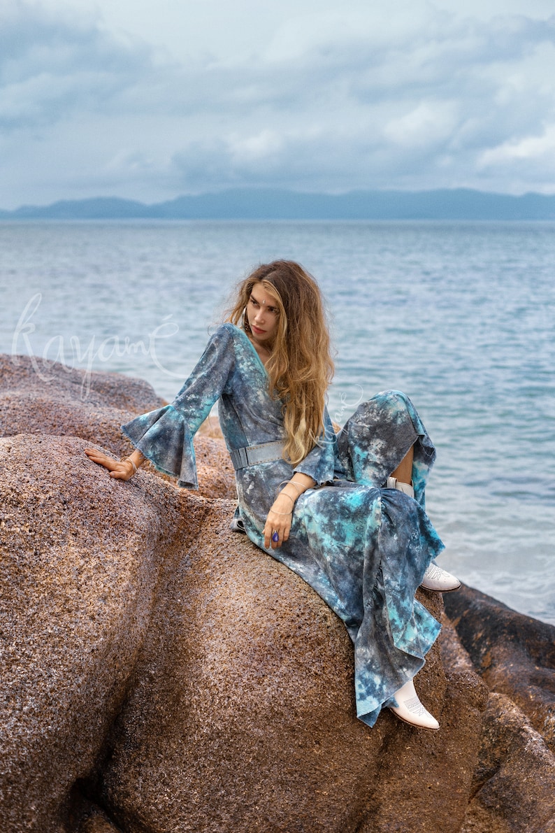 May include: A woman wearing a blue and white tie-dye maxi dress with bell sleeves sits on a large rock by the ocean. She is wearing white boots and has long, wavy hair.