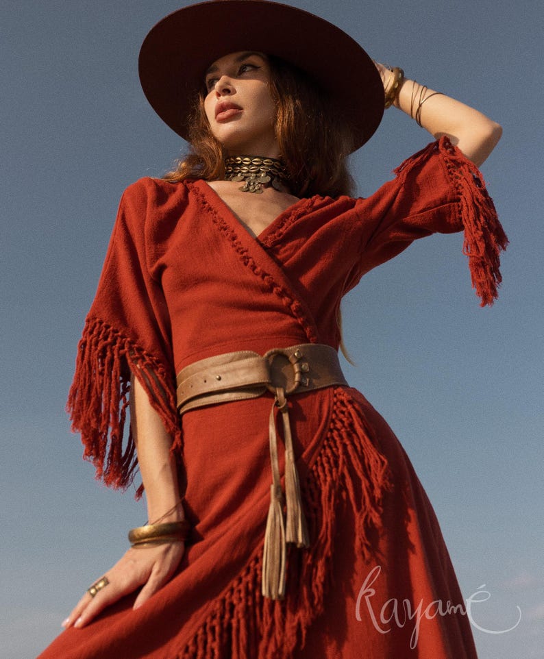 Puede incluir: Un vestido cruzado de color &oacute;xido con flecos en las mangas y el dobladillo. El vestido se ci&ntilde;e a la cintura con un cintur&oacute;n de cuero marr&oacute;n. Un sombrero de ala ancha y joyas completan el look bohemio. El vestido tiene un escote en V.