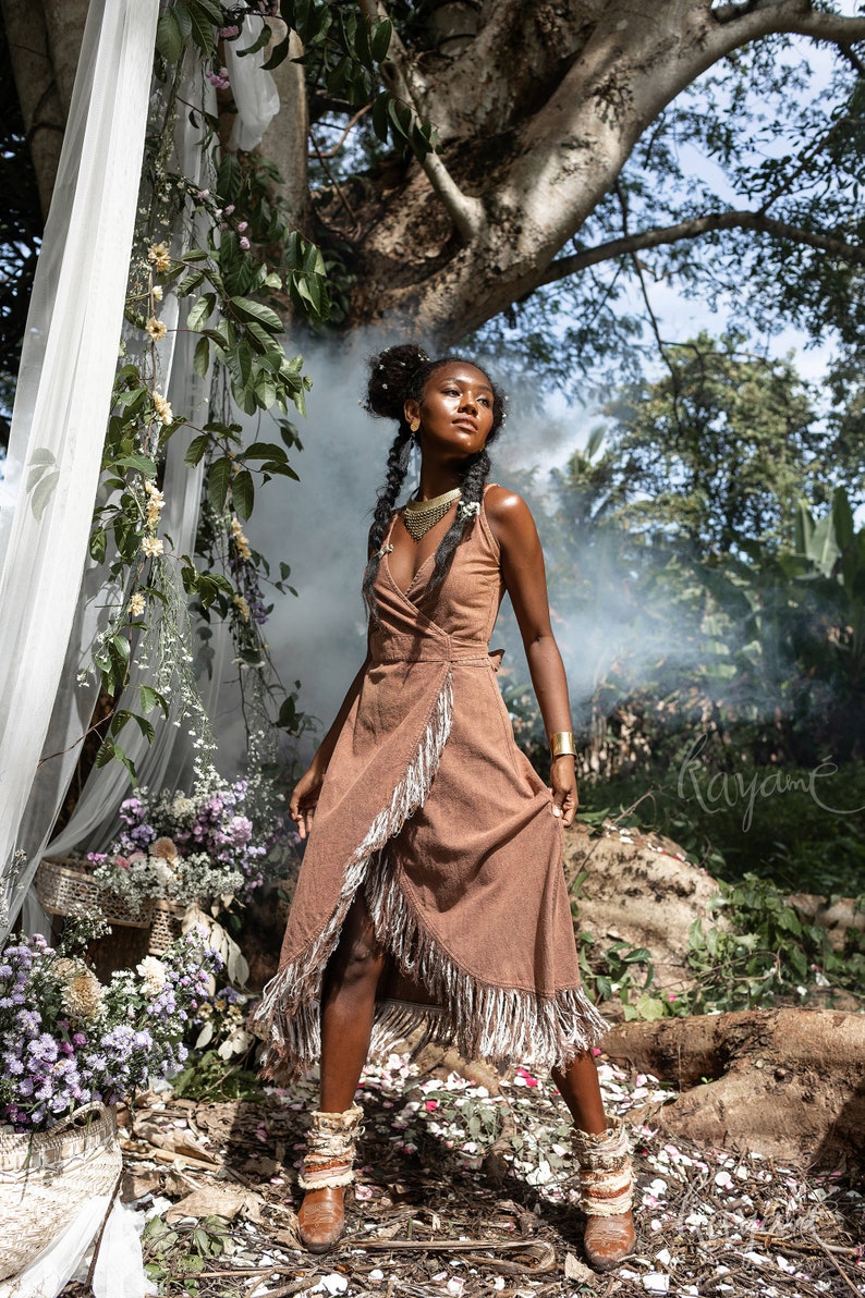 May include: A woman wearing a brown wrap dress with fringe details stands in a forest setting. She is wearing gold jewelry and has her hair in braids. The background features lush greenery and a white sheer curtain.
