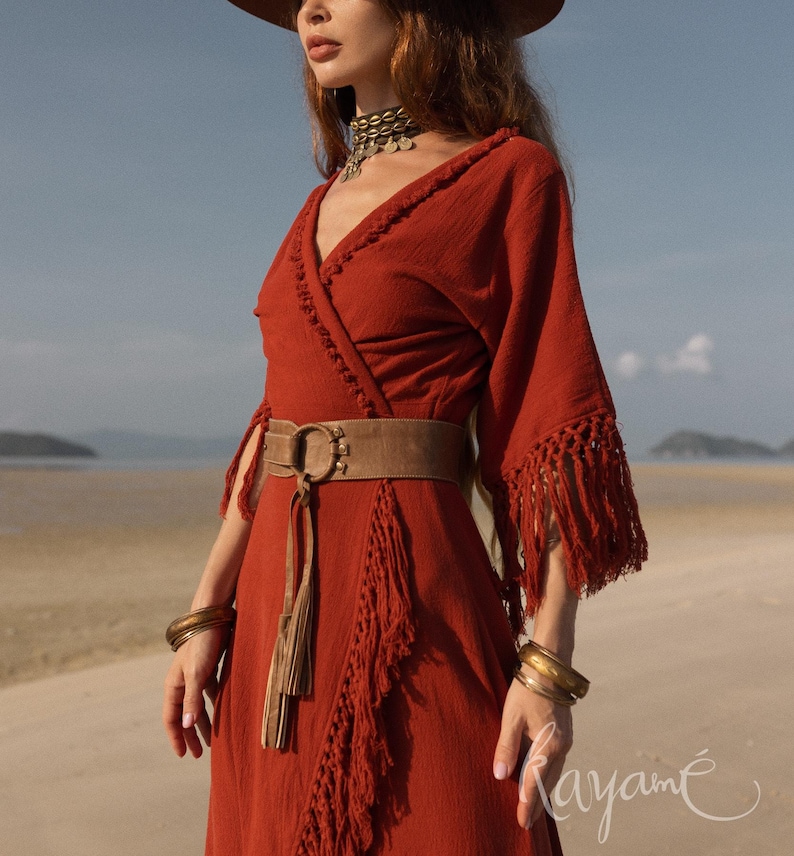 Puede incluir: Un vestido cruzado de color &oacute;xido con flecos en las mangas y la falda. El vestido se ci&ntilde;e a la cintura con un cintur&oacute;n marr&oacute;n con una hebilla circular grande. Brazaletes dorados y un collar gargantilla completan el look bohemio.