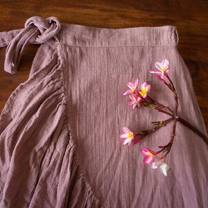 May include: A dusty rose wrap skirt with a gathered ruffle detail and a knotted tie at the waist. A sprig of pink flowers with white centres is lying on the skirt.