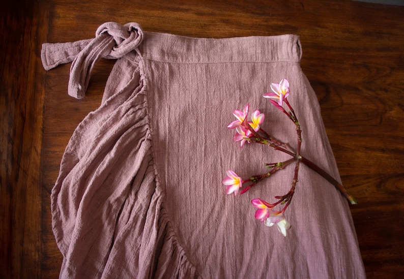 May include: A dusty rose wrap skirt with a gathered ruffle detail and a knotted tie at the waist. A sprig of pink flowers with white centres is lying on the skirt.