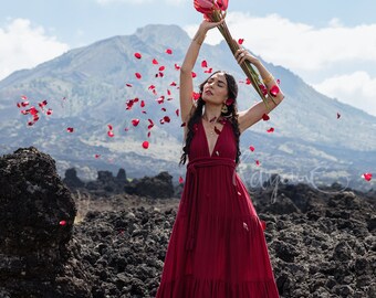 Puede incluir: Una mujer con un vestido maxi rojo se encuentra en un campo de roca volcánica negra, sosteniendo un ramo de flores rojas por encima de su cabeza. Pétalos de rosa caen a su alrededor.