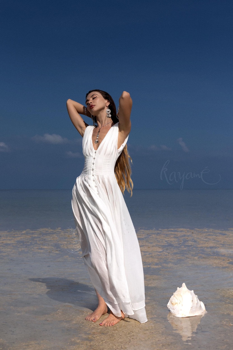 May include: A white, flowing dress with a V-neck and button details. The dress is worn on a beach with a large seashell in the foreground. The sky is blue with a few clouds. The word "Rayane" is written in cursive.