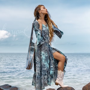 May include: A woman wearing a blue and white tie-dye dress and white cowboy boots stands on rocks by the sea. She is holding a guitar behind her back.