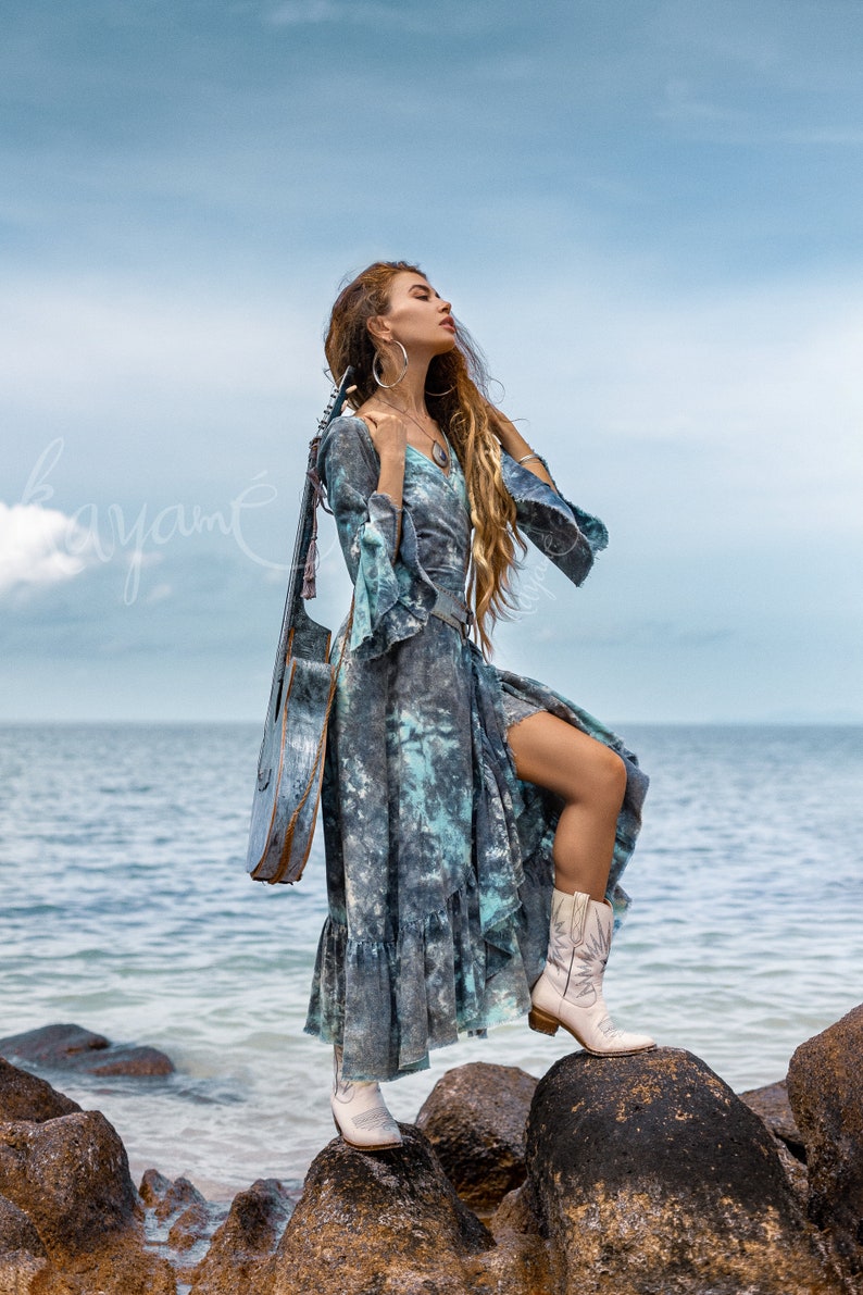 May include: A woman wearing a blue and white tie-dye dress and white cowboy boots stands on rocks by the sea. She is holding a guitar behind her back.