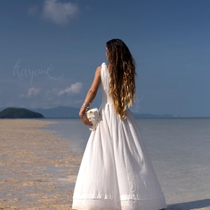 May include: A white sleeveless dress with a flowing skirt is worn by a person standing in shallow water. The person is holding a white object. Long, wavy brown hair cascades down the person's back. The sky is blue with a few clouds. The word "kayane" is written in cursive.
