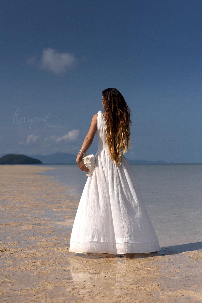 May include: A white sleeveless dress with a flowing skirt is worn by a person standing in shallow water. The person is holding a white object. Long, wavy brown hair cascades down the person's back. The sky is blue with a few clouds. The word "kayane" is written in cursive.