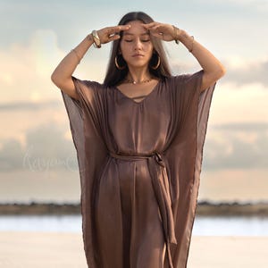 Könnte beinhalten: Fließendes, braunes Seidenkleid mit V-Ausschnitt und passendem Gürtel. Das Kleid hat weite Ärmel und wird mit goldenem Schmuck getragen, darunter Armbänder und Creolen. Der Hintergrund zeigt einen Strand und einen bewölkten Himmel.