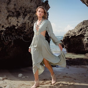 Puede incluir: Una mujer con un vestido maxi verde claro de manga larga y un detalle de corte en la cintura. Est&aacute; de pie en una playa de arena con grandes rocas en el fondo.
