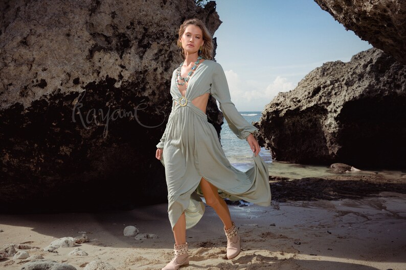 Puede incluir: Una mujer con un vestido maxi verde claro de manga larga y un detalle de corte en la cintura. Est&aacute; de pie en una playa de arena con grandes rocas en el fondo.