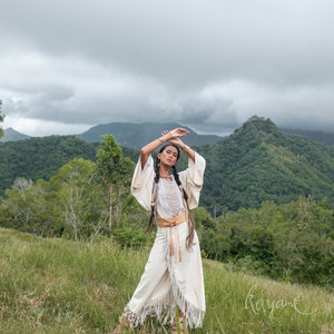 May include: A woman wearing a white, fringed dress with a brown leather belt stands in a grassy field. She has her arms raised above her head and is looking at the camera. The background is a lush green mountain range with a cloudy sky.