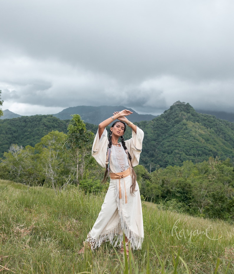May include: A woman wearing a white, fringed dress with a brown leather belt stands in a grassy field. She has her arms raised above her head and is looking at the camera. The background is a lush green mountain range with a cloudy sky.