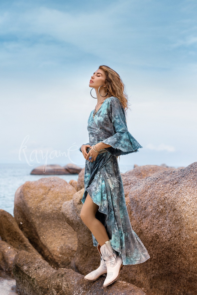 May include: A woman wearing a blue and green tie-dye maxi dress with a ruffled hem and white cowboy boots. She is standing on a rocky beach with the sea in the background.