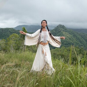 May include: A woman wearing a white, fringed dress with a brown leather belt stands in a field of tall grass. She has long, dark hair and is smiling. The background is a lush green forest with mountains in the distance.